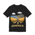 A black short-sleeve crewneck t-shirt featuring a graphic print of a golden road leading to a coastal city, with the word 'AMERICA' printed in gold below, shown on a plain white background.